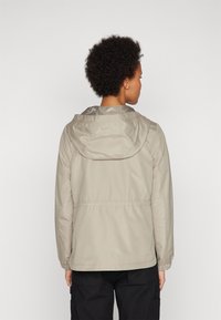 Lyse beige jakke med hette, som har elastiske mansjetter og innsnøring i midjen. Glatt tekstur og minimalistisk design, vist fra baksiden.