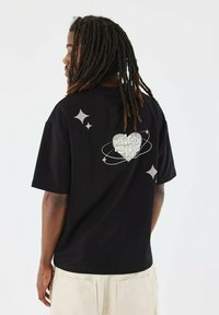 Zwart katoenen T-shirt met oversized pasvorm, voorzien van witte grafische ontwerpen op de achterkant, inclusief tekst en stermotieven. Korte mouwen.