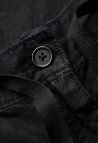 Zwart fabric met een gladde textuur, featuring een ronde zwarte knop met vier gaten en een geweven zwarte band, met opvallende stikdetails.