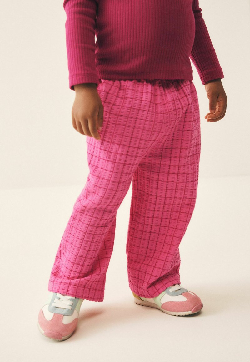 Niño con pantalones rosas a cuadros, una camiseta roja de manga larga y canalé, y zapatillas rosa y blancas, de pie sobre un suelo claro.