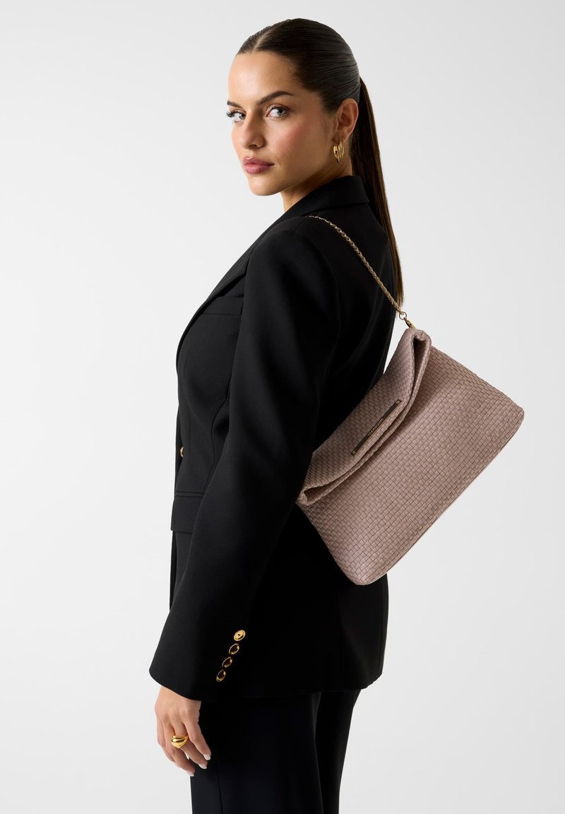 Femme aux cheveux foncés vêtue d'un tailleur noir porte un grand sac à épaule beige texturé avec une sangle chaîne dorée sur un fond blanc uni.