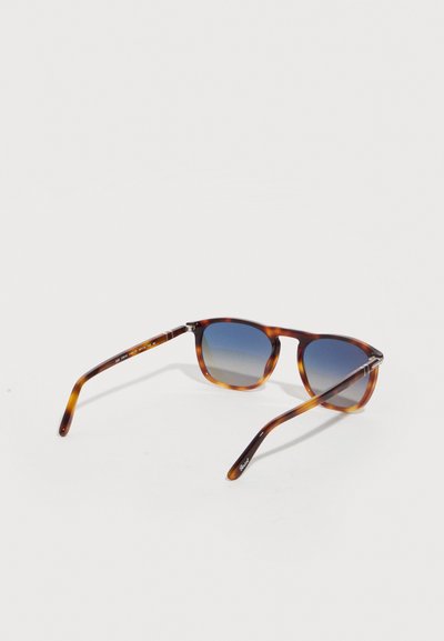 Καφέ γυαλιά ηλίου με σκελετό σε χρώμα χελώνας, με φακούς με μπλε διαβάθμιση, λεπτούς βραχίονες και ελαφρώς τετράγωνο σχήμα, τοποθετημένα σε λευκή επιφάνεια.