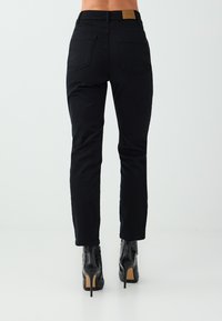 Achteraanzicht van een persoon die zwarte high-waist straight-leg jeans en zwarte enkellaarzen met hoge hakken draagt tegen een effen achtergrond.