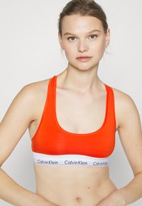 Fényes narancssárga sportmelltartó széles fehér pánttal, amelyen a "Calvin Klein" logo található, sima textúrával és kerek dekoltázsú kialakítással.