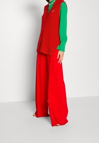 Haut sans manches rouge tricoté porté sur un chemisier vert à manches longues, accompagné d'un pantalon rouge ample. Texture lisse, design simple, look superposé.