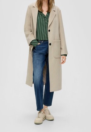 Vrouw die een beige lange jas draagt, een groen gestreept overhemd met knopen, blauwe spijkerbroek en beige sneakers, staand met één hand in haar zak.