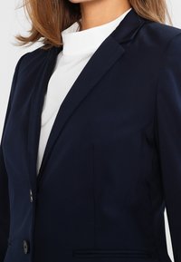 Granatowa marynarka o dopasowanym kroju, jednorzędowa, z dwoma przednimi guzikami i gładką fakturą. Noszona na białej bluzce z wysokim kołnierzem.