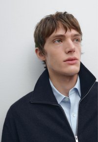 Tmavě modrý zipový svetr s vysokým límcem, vrstvený přes světle modrou košili s knoflíky. Hladká textura, minimální detaily.