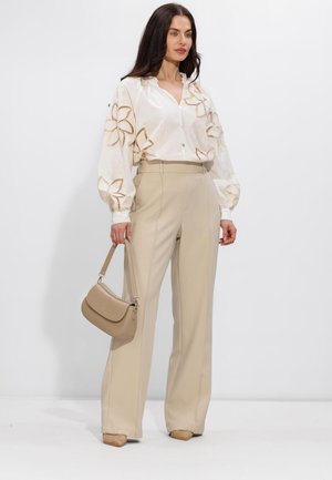 Vrouw draagt een witte blouse met bloemenuitsnijdingen, beige wijde broek, tan schoenen, en houdt een beige handtas vast tegen een witte achtergrond.