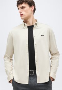 Jeune homme portant une chemise beige à boutons par-dessus un t-shirt noir, avec le logo "BOSS" sur la poitrine, les mains dans les poches, regardant sur le côté.