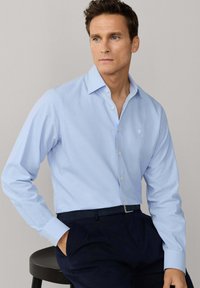 Muž v světle modré košili na knoflíky a tmavě modrých kalhotách sedí na černé stoličce a dívá se mírně doprava.