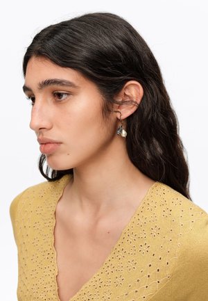 Joven mujer con cabello largo y oscuro que lleva una blusa amarilla bordada y pendientes de plata con detalle de perla, mirando hacia la izquierda de perfil.