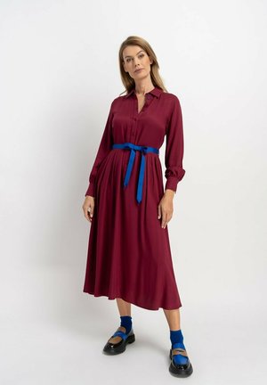 Bordeauxrotes Kleid mit langen Ärmeln, Knöpfen und Kragen, gebunden mit einem blauen Gürtel, kombiniert mit blauen Socken und schwarzen Schuhen. Glatte Stofftextur.