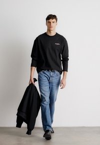 Svart langermet skjorte med logo, blå jeans og svarte sko. Modellen holder en svart jakke. Enkel, ren design på en enkel bakgrunn.