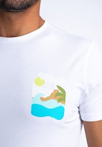 Hvit T-skjorte med et fargerikt trykt design av bølger, en sandstrand, palmeblader, en sol og skyer på venstre brystområde.
