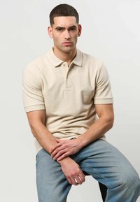 Homme aux cheveux courts et foncés portant un polo beige et un jean bleu clair, assis avec les mains jointes sur ses genoux.