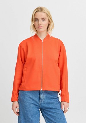 Helle orangene Zip-Jacke aus weichem Stoff, mit Stehkragen, zwei seitlichen Taschen und einem kastigen Schnitt, kombiniert mit blauen Jeans.