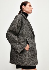 Cappotto a spina di pesce bianco e nero con texture, colletto ampio, chiusura a doppio bottone e tasche laterali; presenta una silhouette morbida e strutturata.