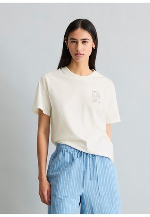 Młoda kobieta z długimi ciemnymi włosami, ubrana w białą koszulkę z nadrukiem i jasnoniebieskie paskowane spodnie sznurkowe, stojąca na jasnoszarym tle.