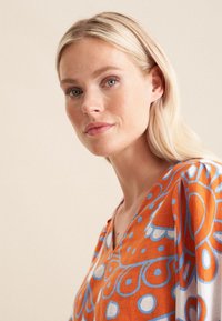 Blusa V-collo colorata in arancione con motivi circolari e floreali blu. Tessuto leggero con una vestibilità rilassata, con maniche corte.