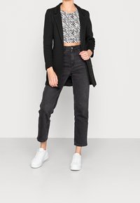 Blazer nero lungo sopra un crop top con stampa zebra, abbinato a jeans neri a vita alta e sneakers bianche. Tessuto liscio e vestibilità su misura.