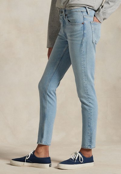 Žmogus dėvi šviesiai mėlynas siauras džinsus, pilką ilgomis rankovėmis palaidinę ir mėlynas drobines sportbačius su baltais raišteliais, stovintis su viena ranka kišenėje.