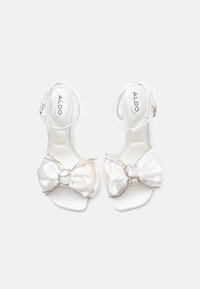 Biele saténové otvorené sandále na vysokých opätkoch s členkovými popruhmi a veľkými ozdobnými mašľami posiatymi rhinestones, umiestnené vedľa seba na bielom pozadí.