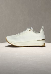 Άσπρο πλεκτό χαμηλό αθλητικό παπούτσι με μπεζ σόλα, κορδόνια στο μπροστινό μέρος, θηλιά στο πίσω μέρος και μεταλλική χρυσή διακόσμηση στο πίσω μέρος.