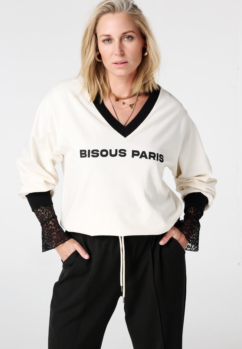 Roomkleurige V-hals sweatshirt met "BISOUS PARIS" in zwarte letters, met zwarte kanten accenten op de mouwen. Gecombineerd met zwarte joggerbroek.