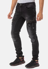 Pantalons cargo en denim noir avec plusieurs poches, zippers et une jambe fuselée. Caractéristiques d'un délavage dégradé et d'accents côtelés près des chevilles.