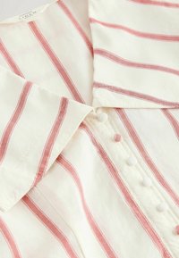 Chemise en lin blanc avec des rayures verticales rouges, boutons recouverts de tissu et col, montrant un gros plan de l'avant et de la manche.