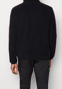 Svart fleecejacka med hög krage, röda sidosträck och ton-i-ton-sömmar. Har långa ärmar och en avslappnad passform.