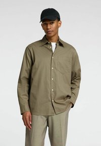 Camicia a maniche lunghe verde oliva, in cotone, con bottoni sul davanti, taschino sul petto, abbinata a pantaloni grigi chiari e un berretto nero.