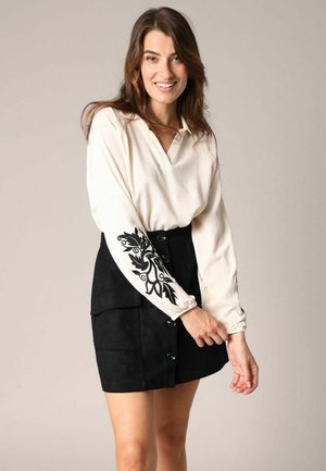 Chemise blanche à manches longues en tissu texturé avec broderie florale noire sur les manches, associée à une jupe noire en suède à boutons sur le devant.