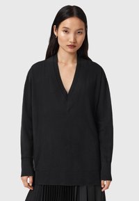 Jonge vrouw met lang, donker haar die een oversized zwarte V-halstrui en een geplooide zwarte rok draagt, staande tegen een effen grijze achtergrond.