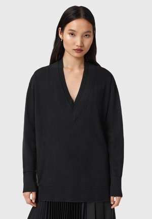 Jeune femme aux longs cheveux noirs portant un pull noir oversized à col en V et une jupe plissée noire, se tenant devant un fond gris uni.