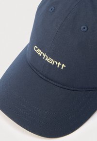 Μπλε σκούρο μπέιζμπολ καπέλο από υφασμα με υφή. Διαθέτει κεντημένο λογότυπο "carhartt" σε ανοιχτό κίτρινο και ραμμένη λεπτομέρεια στην περούκα.