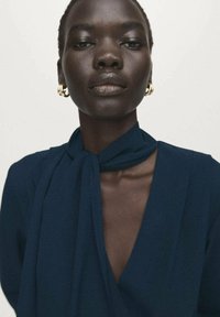 Marineblauwe blouse met striksluiting, voorzien van een diep V-hals, vloeiende stof en subtiele plooien. Model draagt gouden oorringen.