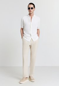 Camicetta bianca a maniche corte con finitura testurizzata, pantaloni beige e scarpe chiare, abbinate a occhiali da sole scuri.