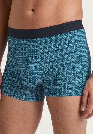 Männliche Boxer-Shorts aus einem türkisfarbenen Stoff mit geometrischem Muster. Verfügt über einen marineblauen elastischen Bund mit eingeprägtem Markennamen.