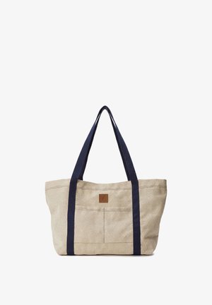 Beżowa torba na ramię z płótna z granatowymi paskami, ozdobiona przeszyciami oraz kwadratową, brązową skórzaną metką z przodu.