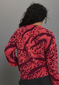 Strickpullover in leuchtendem Koralle mit dunkellila abstrakten Mustern. Hat einen gerippten Saum und lange Ärmel und bietet eine lockere Passform.