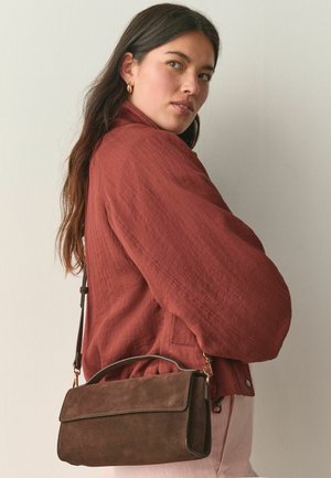 Naine, kes kannab roostevärvi jope ja kuldsete rõngaskõrvarõngastega, kandmas pruuni seemisnahast õlakotti, mis on ühtlase taustaga.