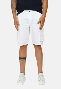 Pantaloni cargo bianchi con due tasche laterali, realizzati in tessuto leggero. Indossati con scarpe da ginnastica bianche e presentano un tatuaggio a strisce nere sopra il ginocchio.