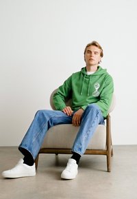 Πράσινη κουκούλα με κορδόνια και γραφικά στοιχεία, συνδυασμένη με ανοιχτό μπλε τζιν και λευκά αθλητικά παπούτσια, καθισμένη σε ξύλινη καρέκλα.