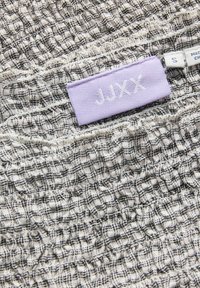 Riietesilt tekstureeritud must-valgel ruudulisel kangal, millel on lavendlivärvi silt tekstiga "JJXX" ja väike valge suurusesilt "S".