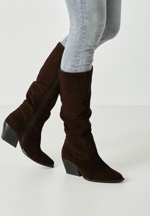 Person trägt dunkelbraune Wildleder-Stiefel bis zur Mitte der Wade mit schwarzen Keilabsätzen und hellgrauen enganliegenden Jeans vor einfarbigem Hintergrund.