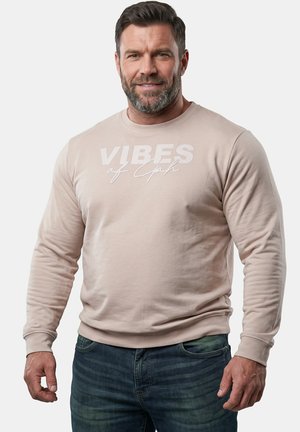Uomo con capelli corti e scuri e barba, che indossa una felpa beige con il testo "VIBES of God" e jeans blu scuro, in piedi davanti a sfondo bianco.