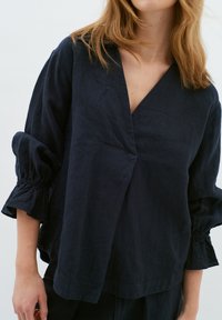 Blusa in lino blu navy con design avvolgente, scollo a V e maniche a palloncino con polsini elasticizzati. Texture liscia, vestibilità comoda.
