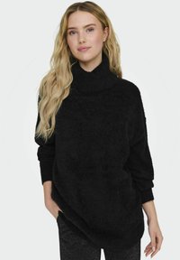 Maglione nero in tessuto morbido con collo a dolce vita, maniche ampie a pipistrello e vestibilità rilassata. Texture soffice e orlo leggermente asimmetrico.